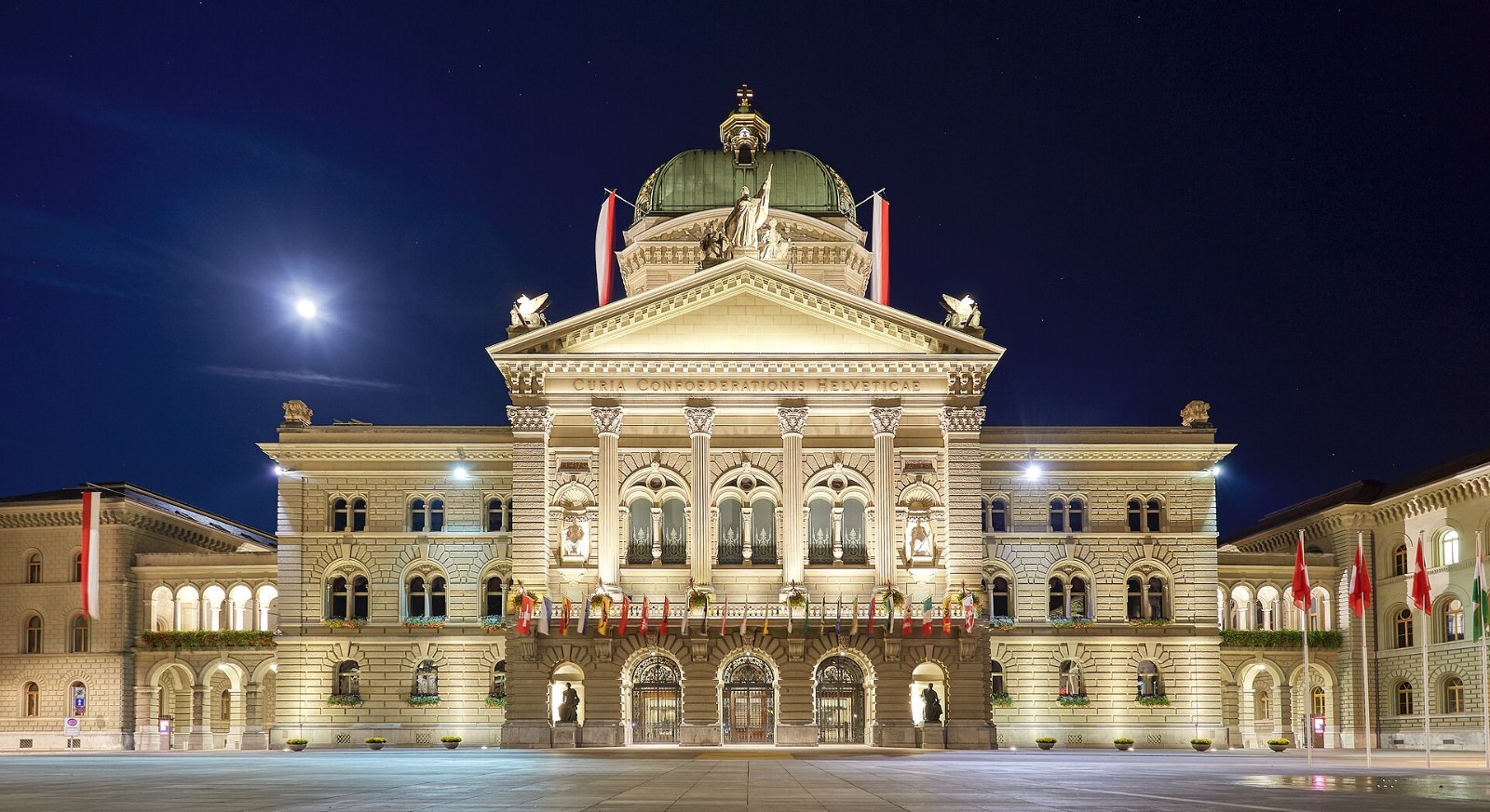 Das beflaggte Bundeshaus am Bundesplatz in Bern bei Nacht